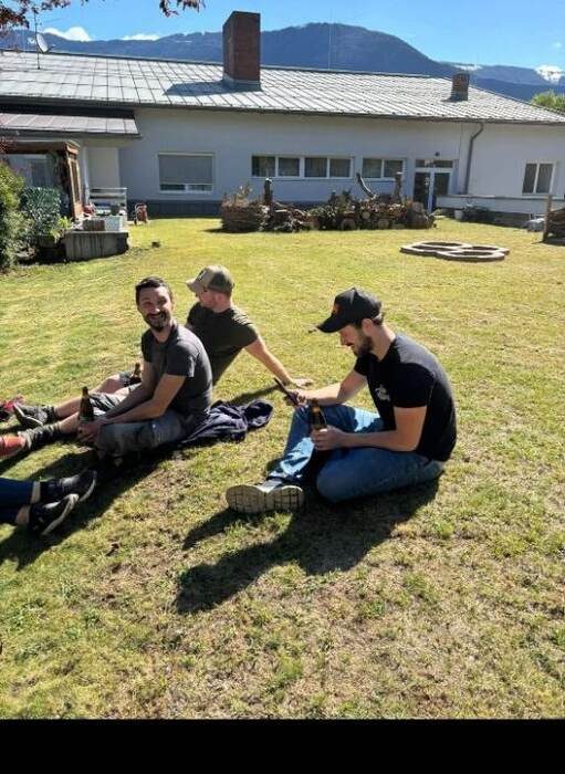 Drei Helfer sitzen nach getaner Arbeit lachend auf der Wiese vor dem Gebäude, im Hintergrund die frisch angelegten Gartenelemente und die Bergkette