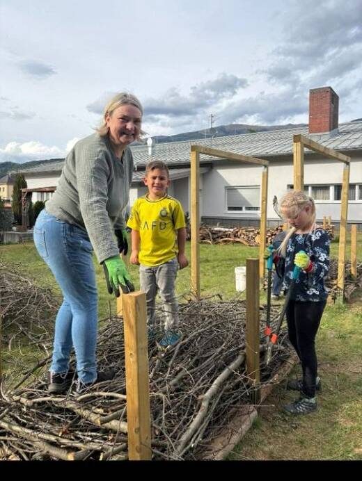 Eine Helferin und zwei Kinder schichten Äste in die Holzumrandung einer Benjeshecke, im Hintergrund die Berge des Drautals